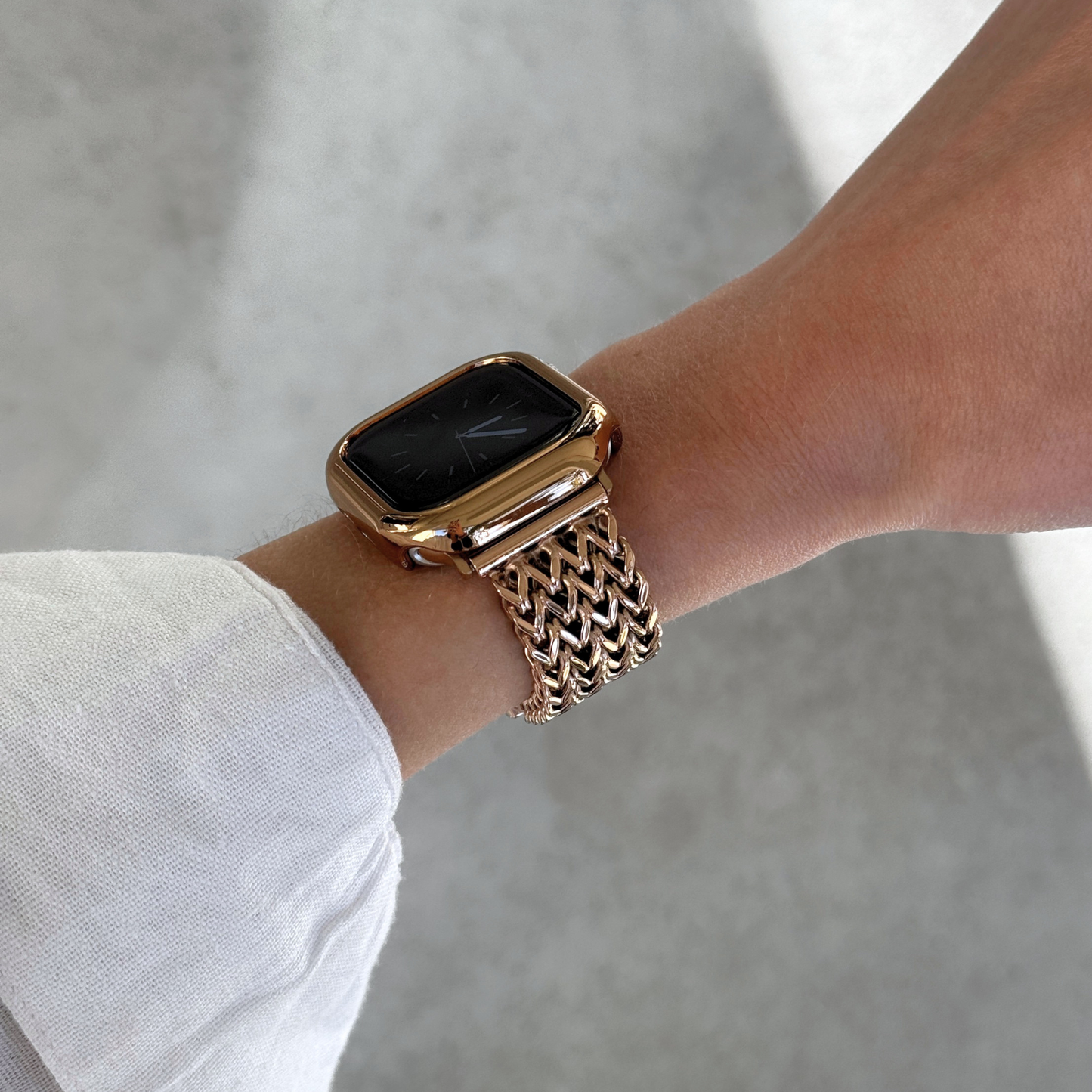 A close-up of a person's wrist wearing a rose gold-toned Apple smartwatch with a matching metallic mesh band and bezel, set against a soft gray concrete background.