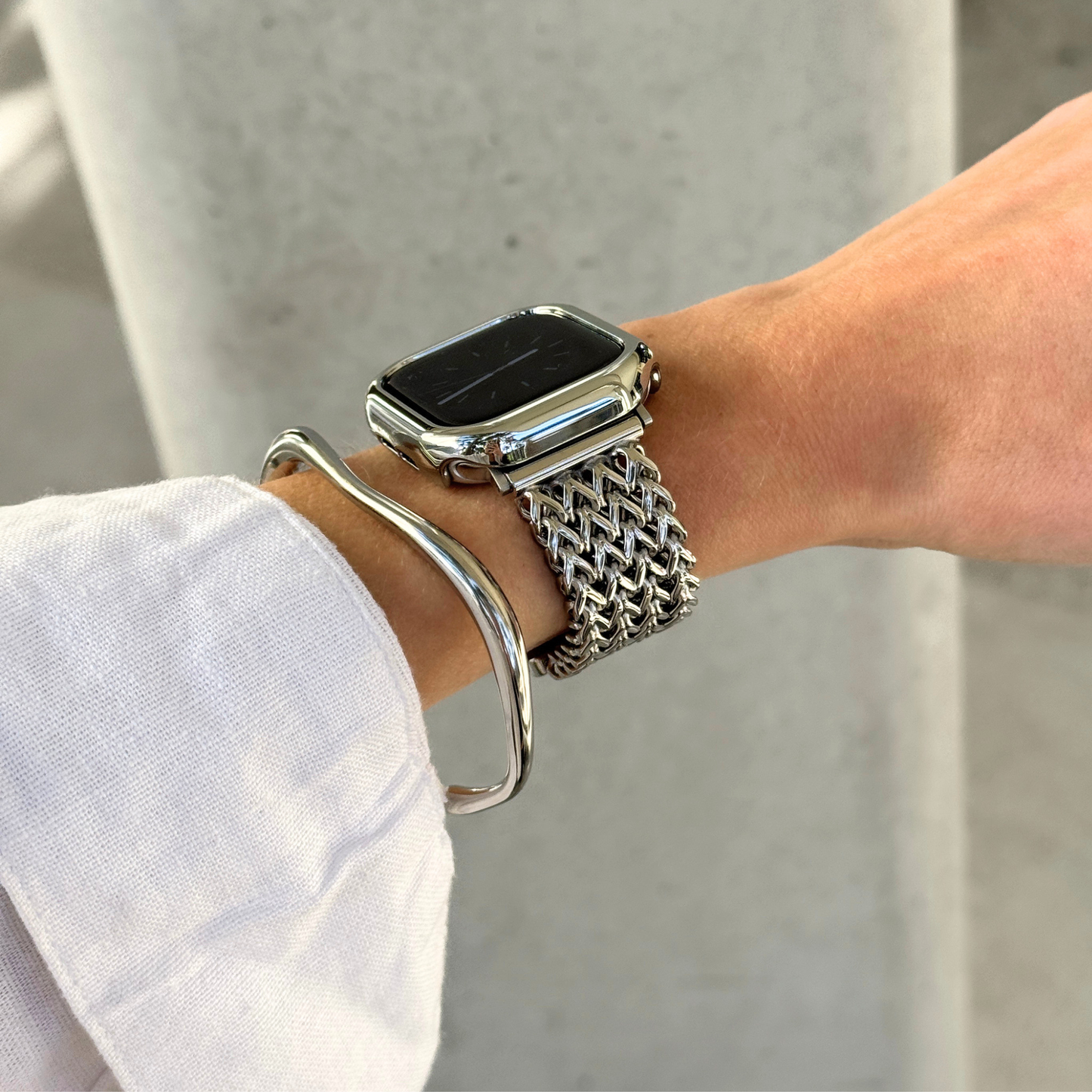 A close-up of a person's wrist wearing a silver mesh Apple watch band with a smartwatch, along with a silver bangle bracelet and a matching bezel, against a light gray concrete background.