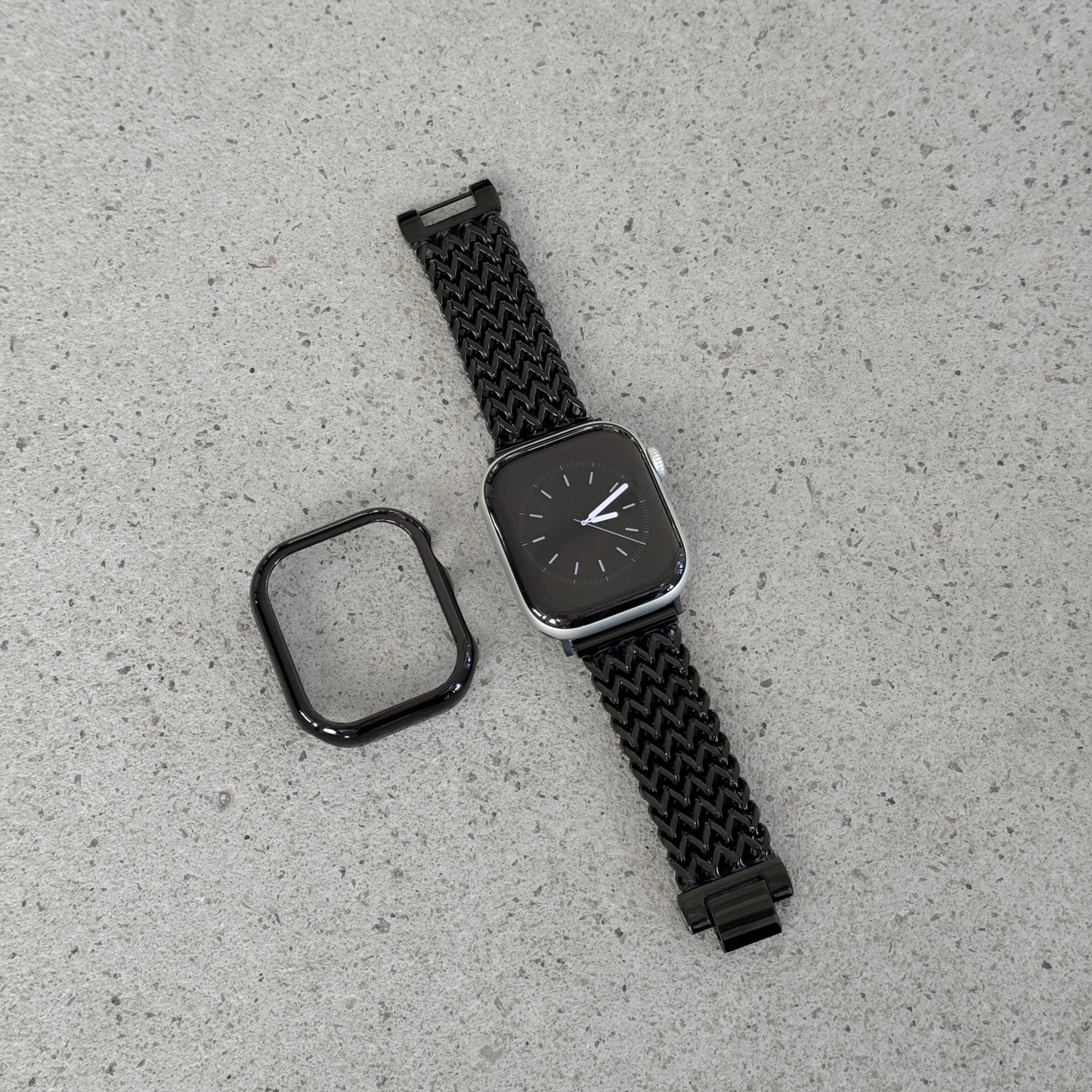 A black smartwatch with a woven black mesh strap and a separate black protective case, displayed on a light gray textured surface.