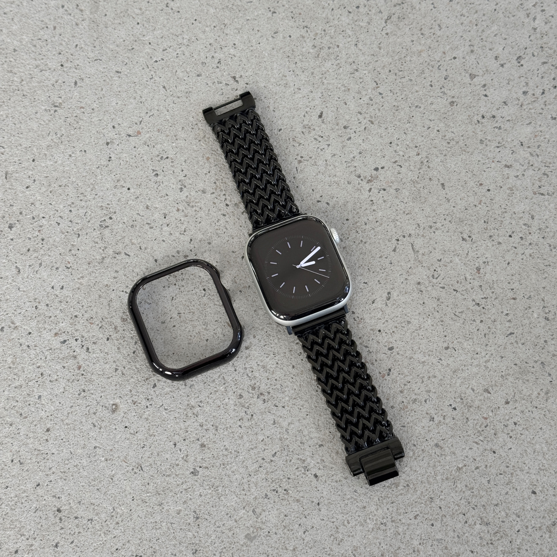 A black smartwatch with a woven black mesh strap and a separate black protective case, displayed on a light gray textured surface.