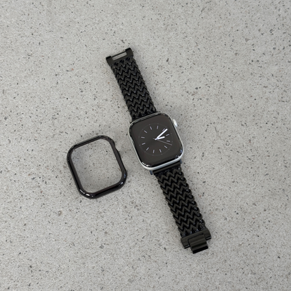 A black smartwatch with a woven black mesh strap and a separate black protective case, displayed on a light gray textured surface.