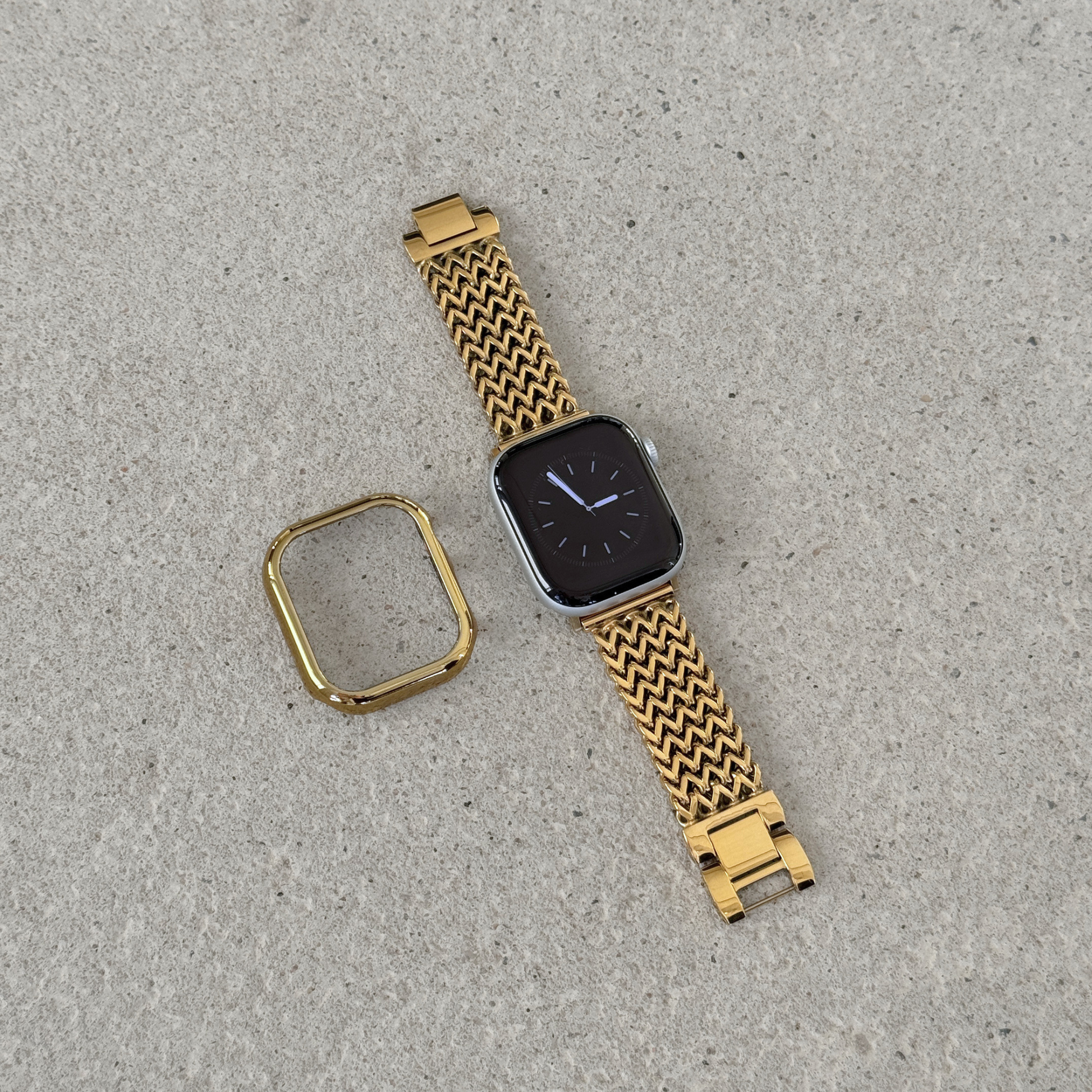 A gold-tone metal mesh Apple watch band with a matching gold bezel protector, shown on a silver smartwatch against a textured gray concrete background.