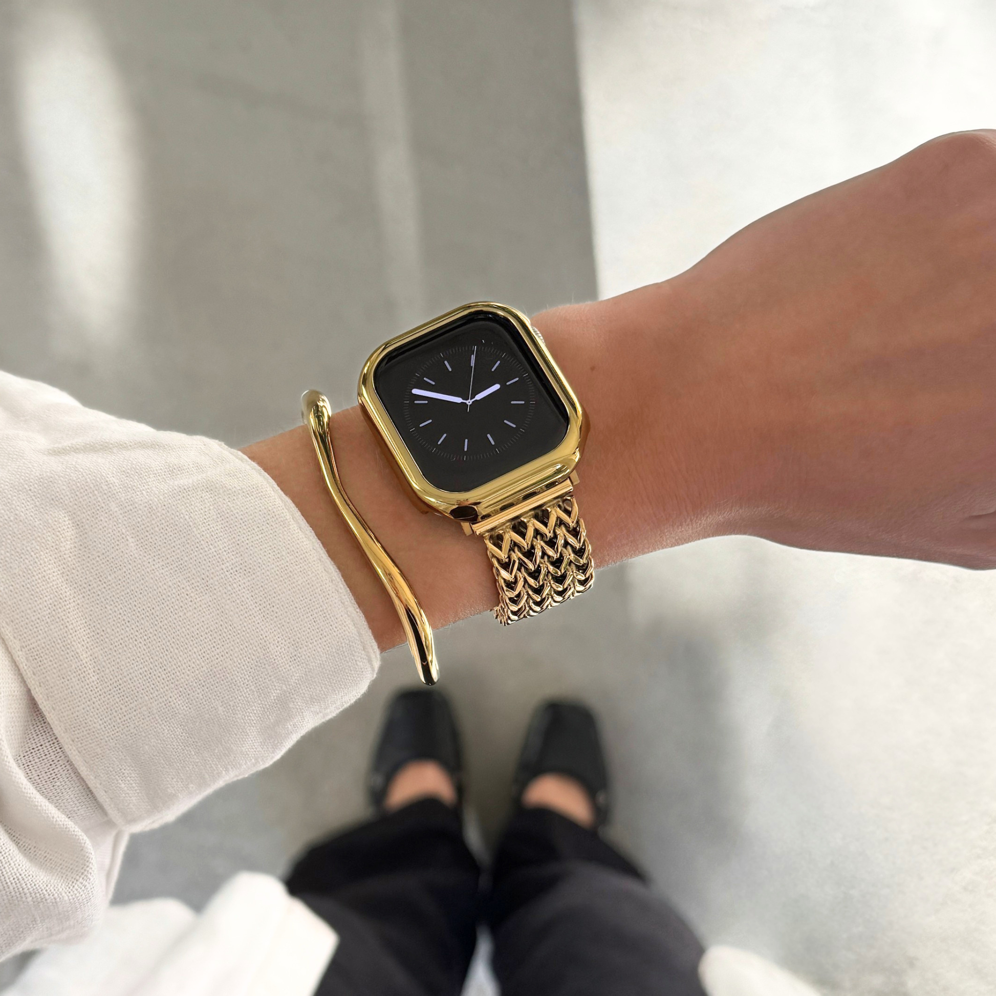 A gold-toned textured Apple Watch mesh band, bundled with a matching gold bezel case and cuff bracelet, worn on a person's wrist. The person is also wearing a white long-sleeve top, with black pants and shoes visible below against a light gray floor.