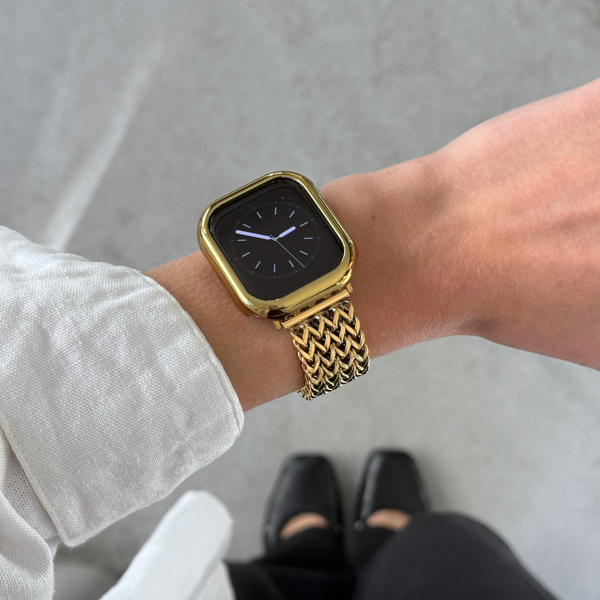 A gold smartwatch with a gold mesh band worn on a person's wrist, shown in a lifestyle setting with a gray concrete floor and black shoes visible.