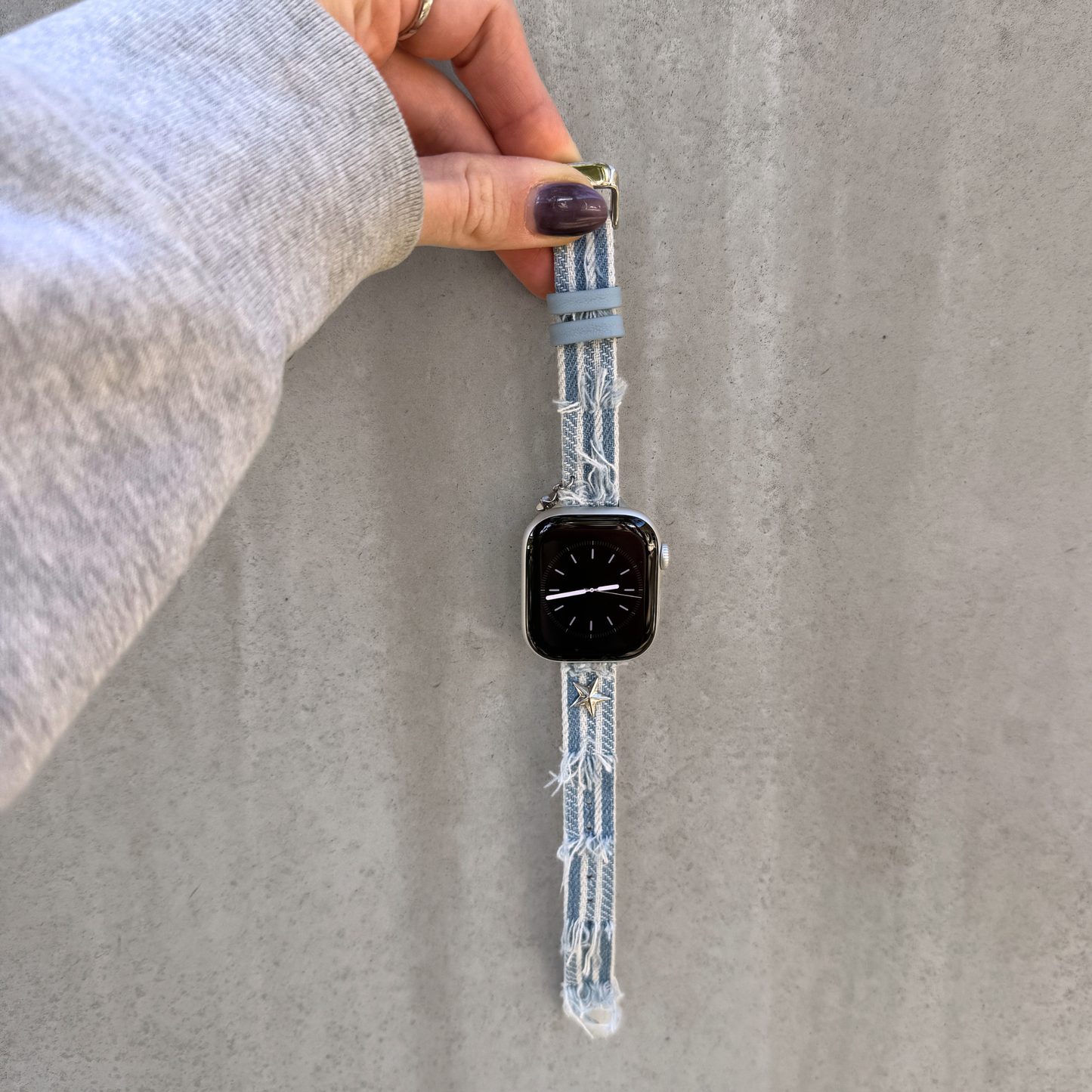 A light blue denim Apple watch strap with frayed edges and a matching light blue loop, shown attached to a smartwatch and held by a hand against a gray concrete wall.