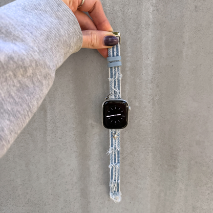 A light blue denim Apple watch strap with frayed edges and a matching light blue loop, shown attached to a smartwatch and held by a hand against a gray concrete wall.