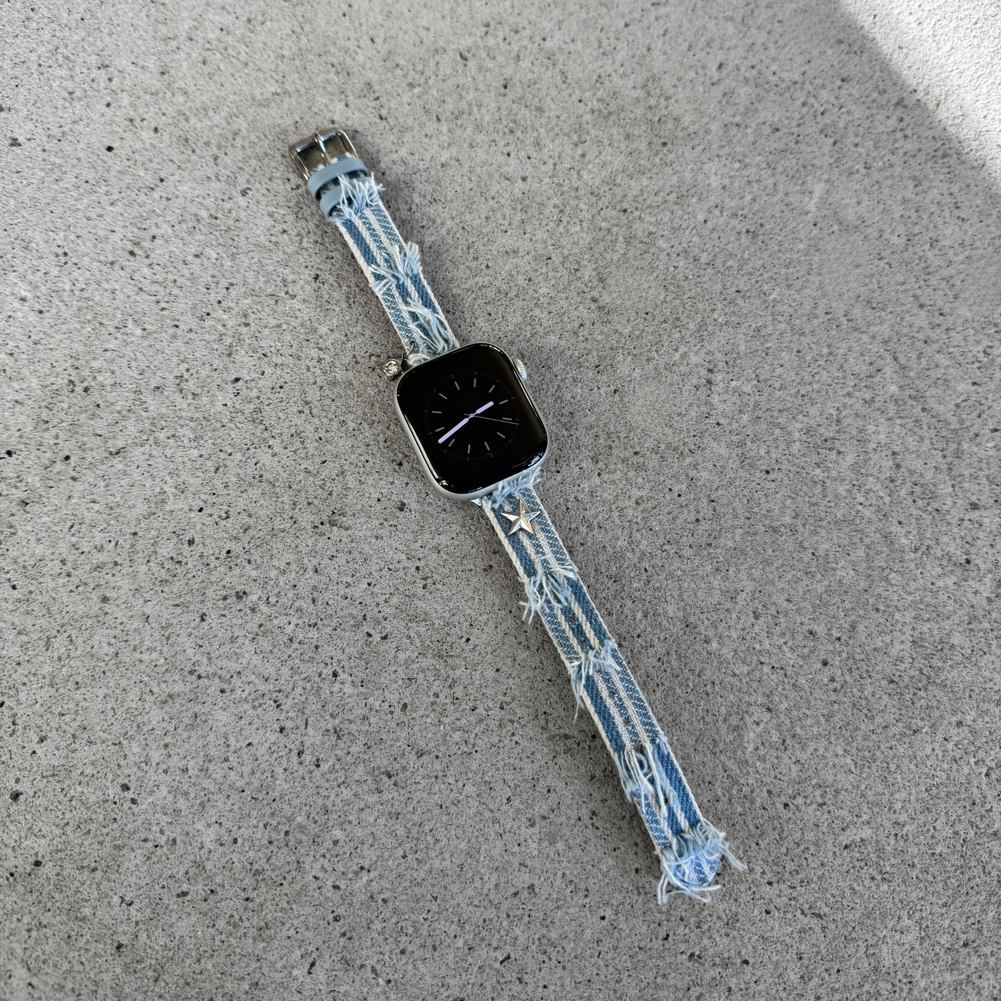 A light blue denim Apple watch strap with frayed edges and small star charms, attached to a smartwatch, lying on a textured gray concrete surface.