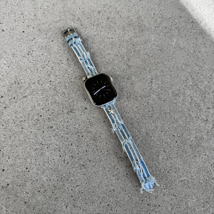 A light blue denim Apple watch strap with frayed edges and small star charms, attached to a smartwatch, lying on a textured gray concrete surface.