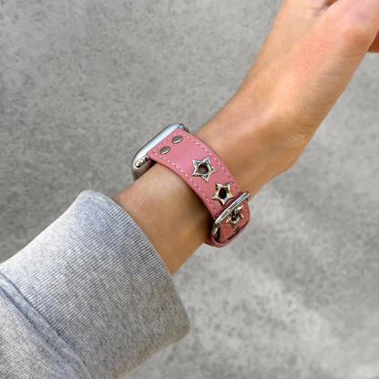 A pink leather Apple watch band with silver-tone metal star-shaped studs, shown worn on a person's wrist with a gray long-sleeve shirt visible.