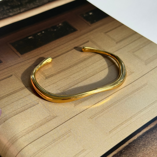 A polished gold-toned open cuff bangle bracelet with a wavy, organic curve, resting on a textured beige surface.