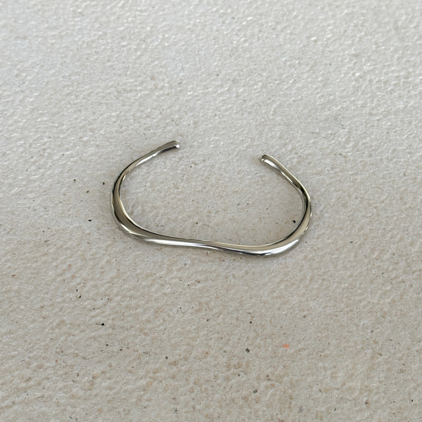 A polished silver-toned metal cuff with a wavy, organic curve, displayed on a textured off-white surface.