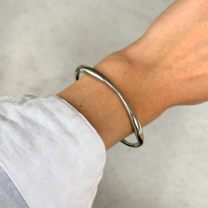 A polished silver-toned metal bangle bracelet worn on a person's wrist, shown against a light gray background with a white shirt cuff visible.