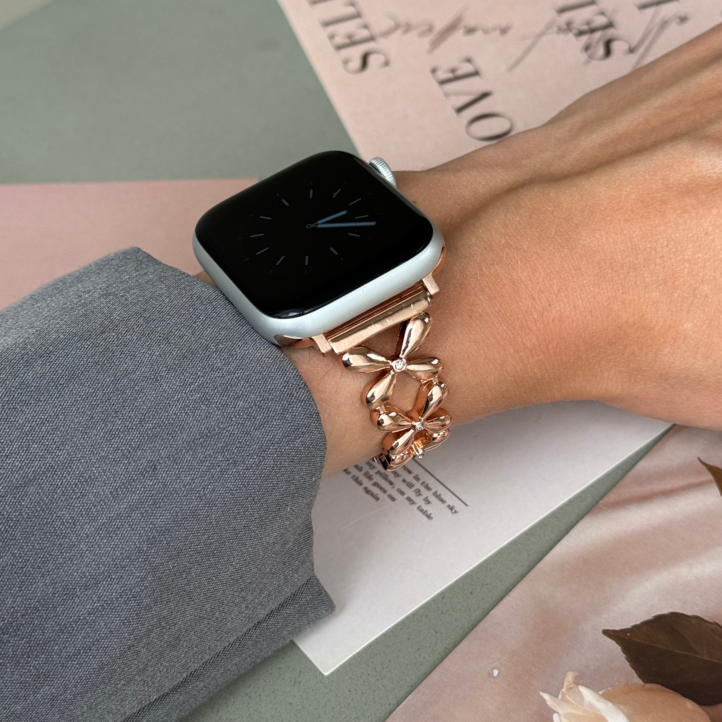 Rose gold floral link Apple Watch band worn on a wrist beside pink paper cards, metal strap for Apple Watch 38mm to 49mm