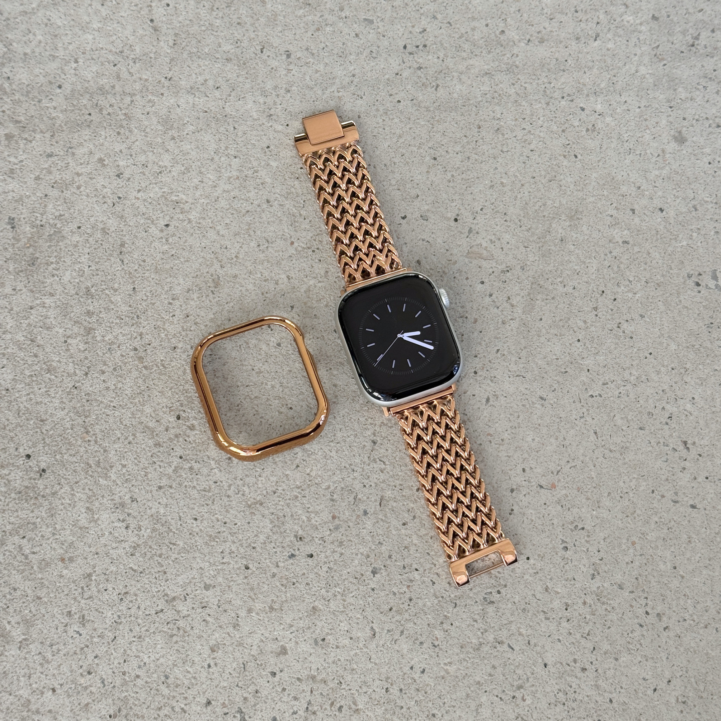 A rose gold metal mesh Apple watch band with a matching square bezel, shown on a smartwatch alongside its detached strap end, placed on a textured gray concrete surface.