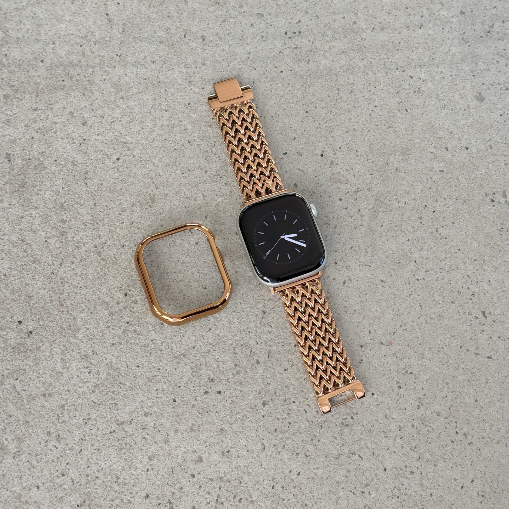 A rose gold metal mesh Apple watch band with a matching square bezel, shown on a smartwatch alongside its detached strap end, placed on a textured gray concrete surface.