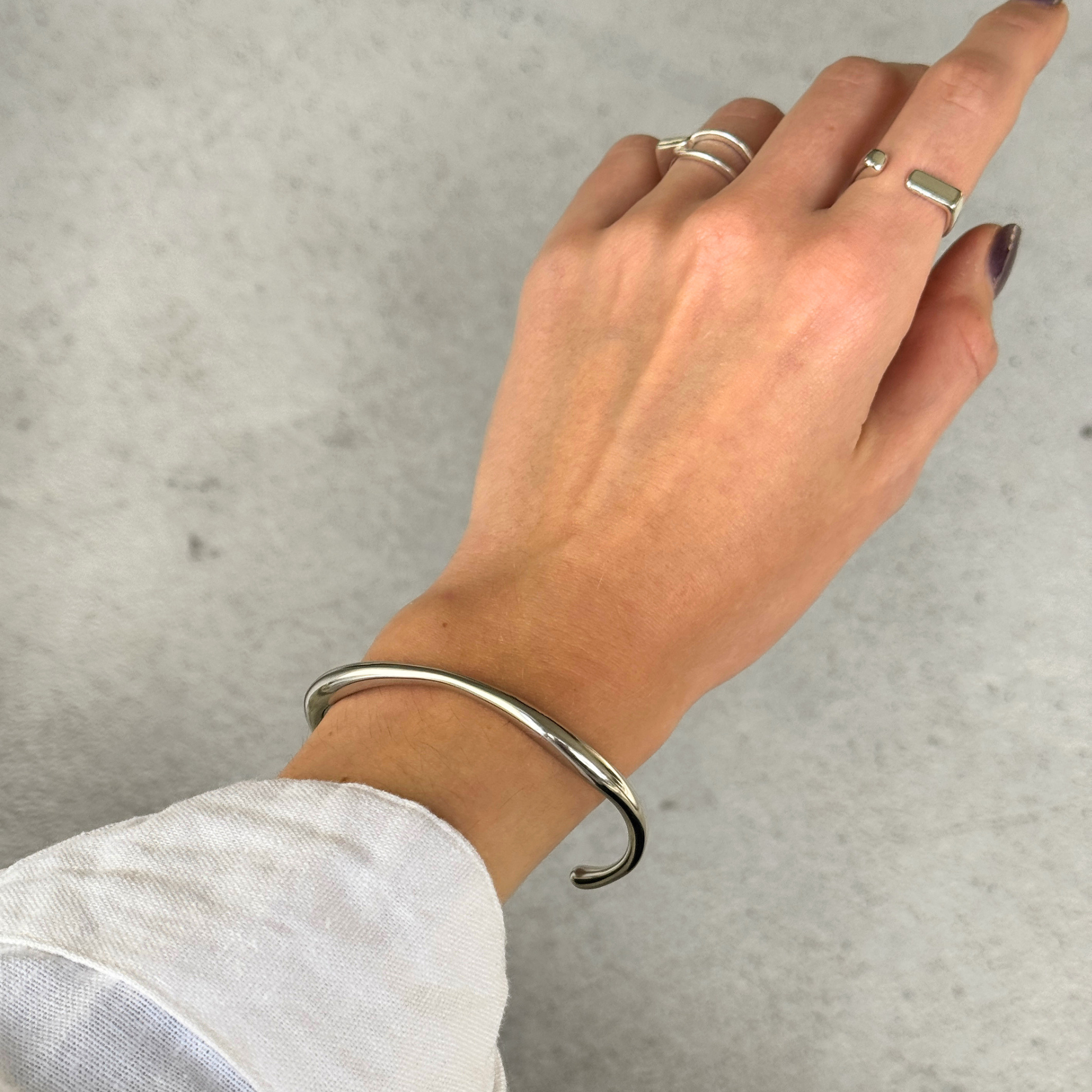 A silver-toned metal hinged cuff bracelet worn on a person's wrist, with their hand resting against a light gray textured background.