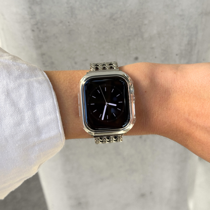 A silver Apple Watch with a clear bumper case with a decorative chain strap worn on a person's wrist, shown against a light gray concrete wall.