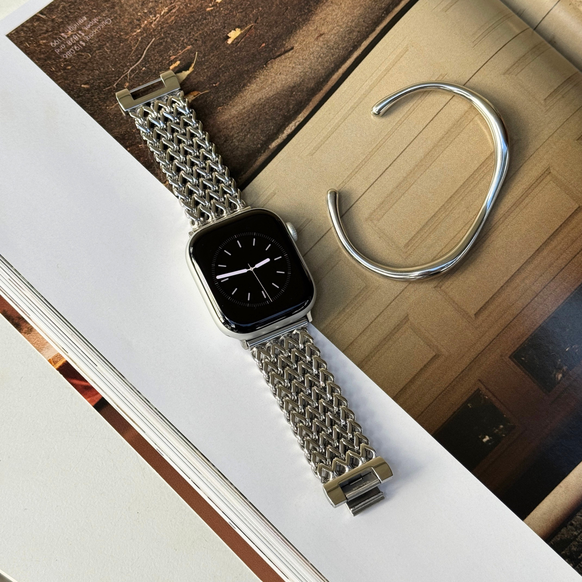 A silver mesh metal watch band attached to an Apple smartwatch with a black display, styled on top of an open book next to a silver cuff bracelet.