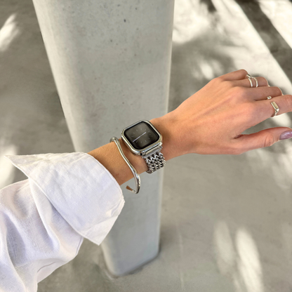 A silver metal Apple watch link strap with a matching silver-toned cuff bracelet and a silver case cover, worn on a person's wrist, styled with a silver bangle and rings, set against a concrete pillar background.
