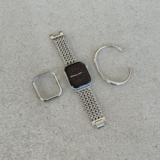 A silver stainless steel mesh Apple watch band with a matching silver bezel protector and a matching cuff bracelet, arranged on a textured gray surface next to an Apple Watch.