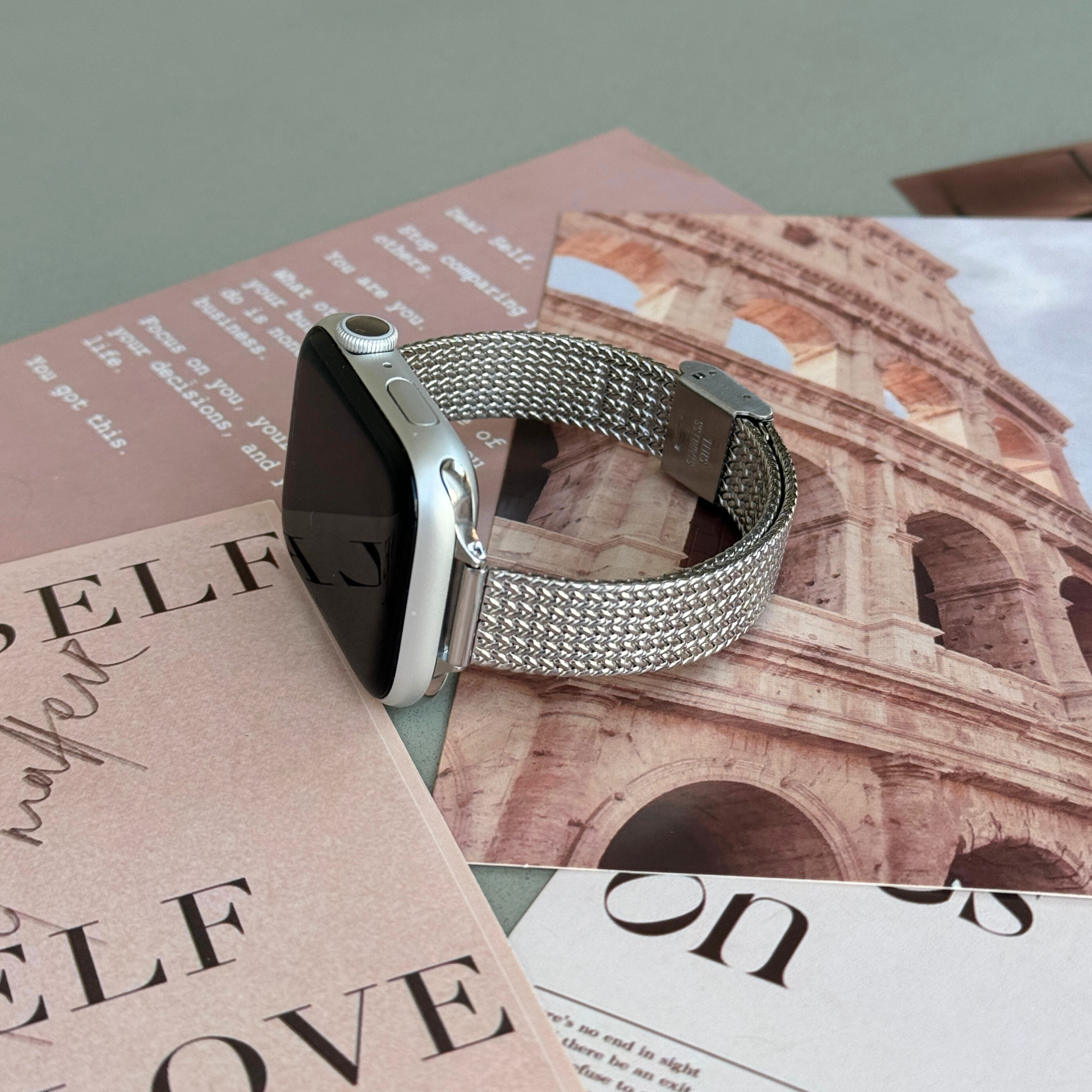Silver woven mesh Apple Watch band lying flat on magazine pages, stainless steel bracelet fitting Apple Watch 38mm to 49mm