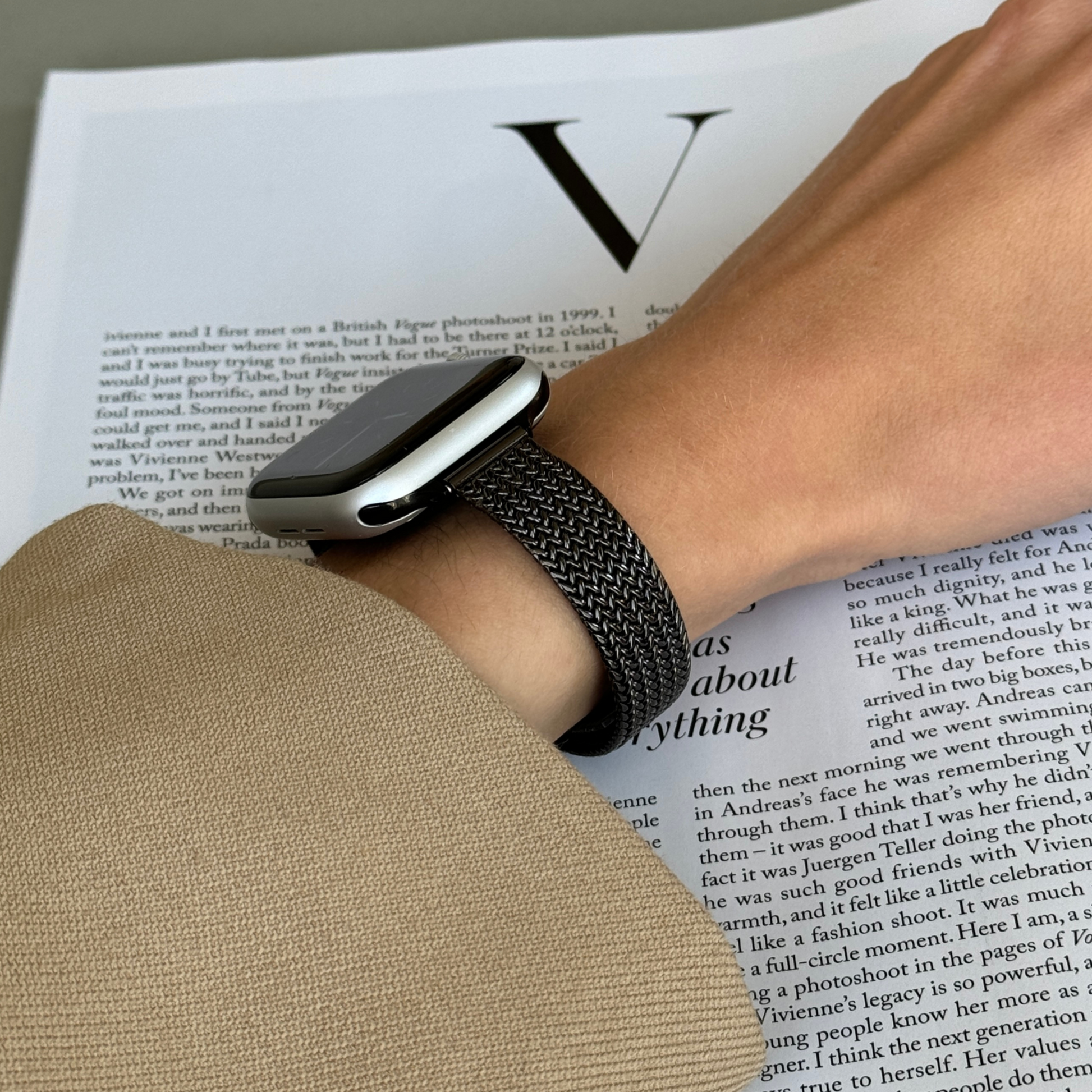 Black woven mesh Apple Watch band worn on a wrist above an open newspaper, stainless steel strap for Apple Watch 38mm to 49mm