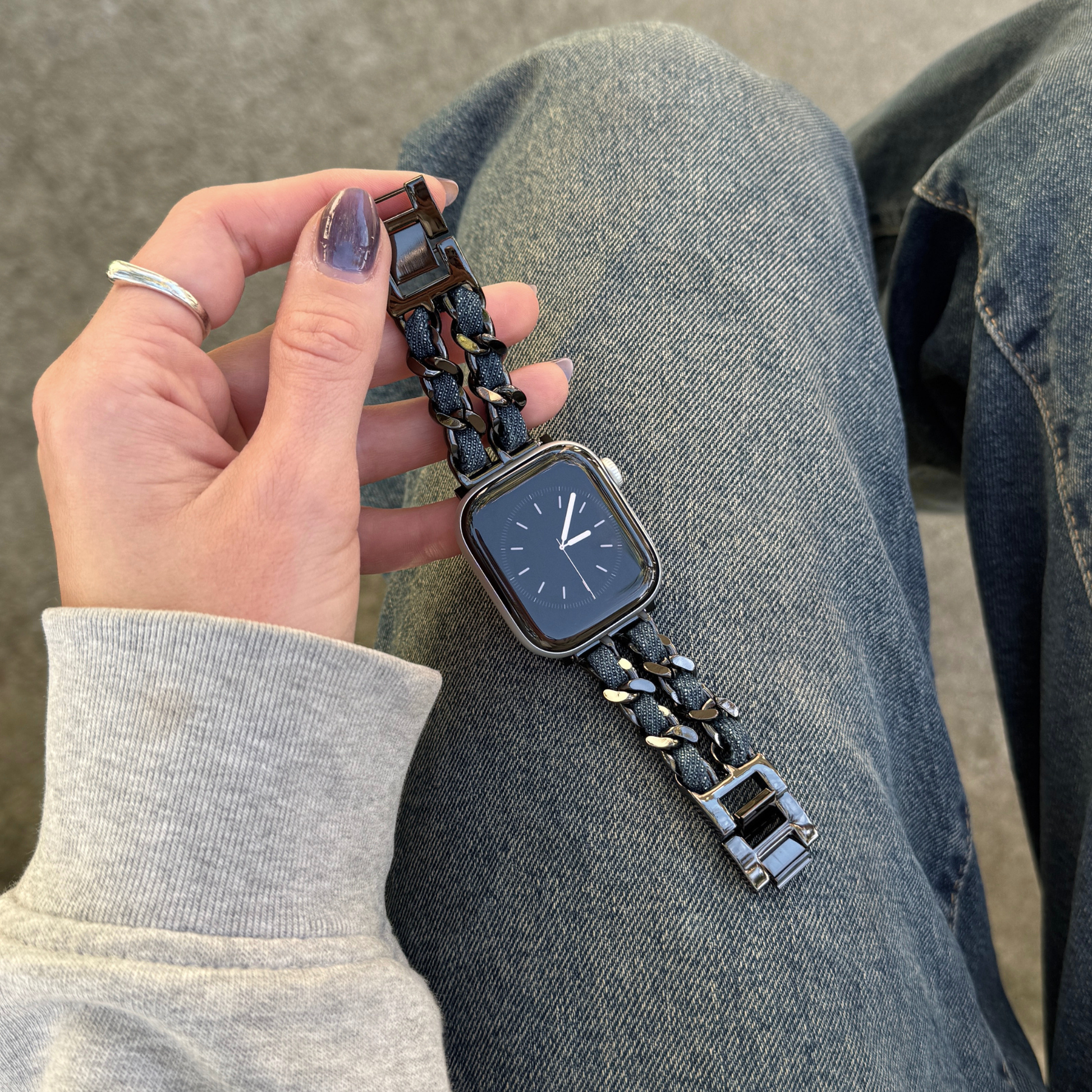 A close-up of a person's hand holding a an Apple Watch with a black metallic link band, set against their denim-clad legs and a gray floor.