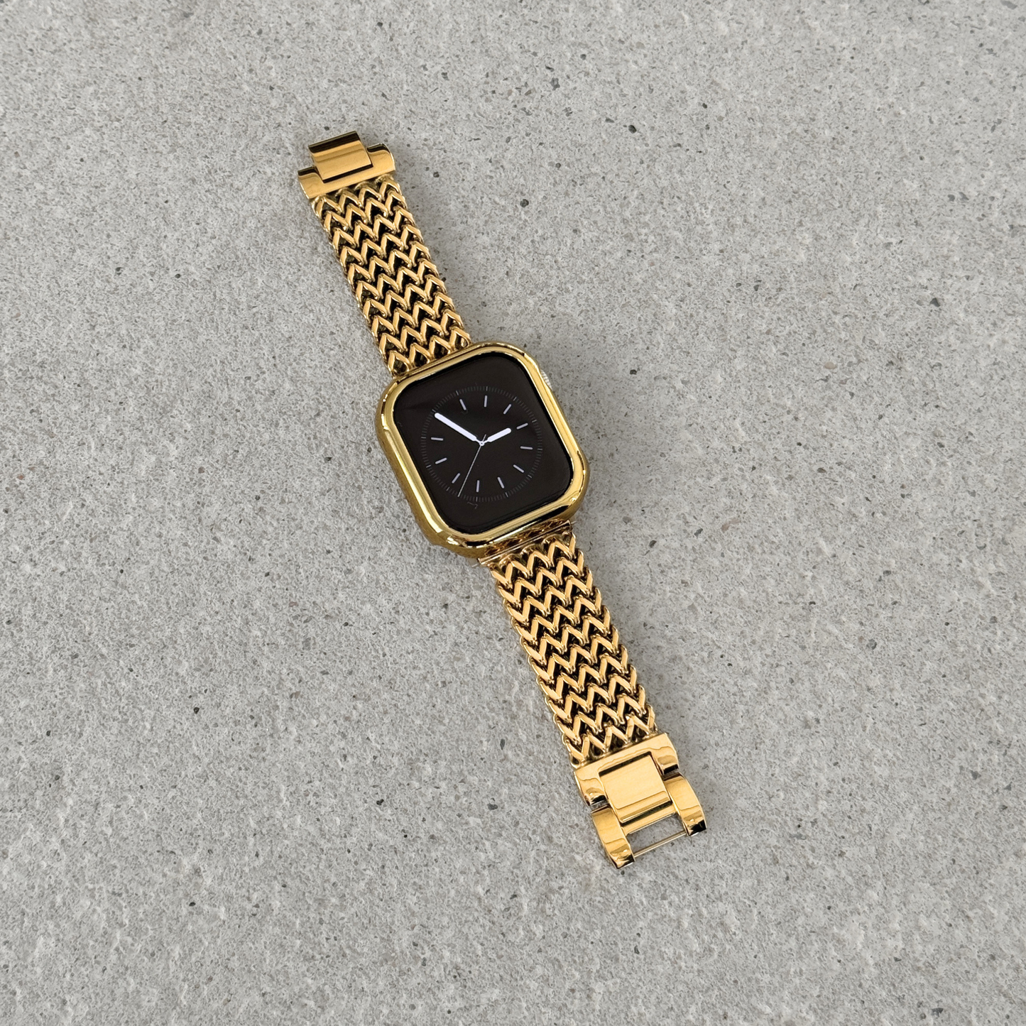 Apple Watch with a gold bumper case cover and a woven metallic mesh strap, photographed diagonally on a textured gray surface.