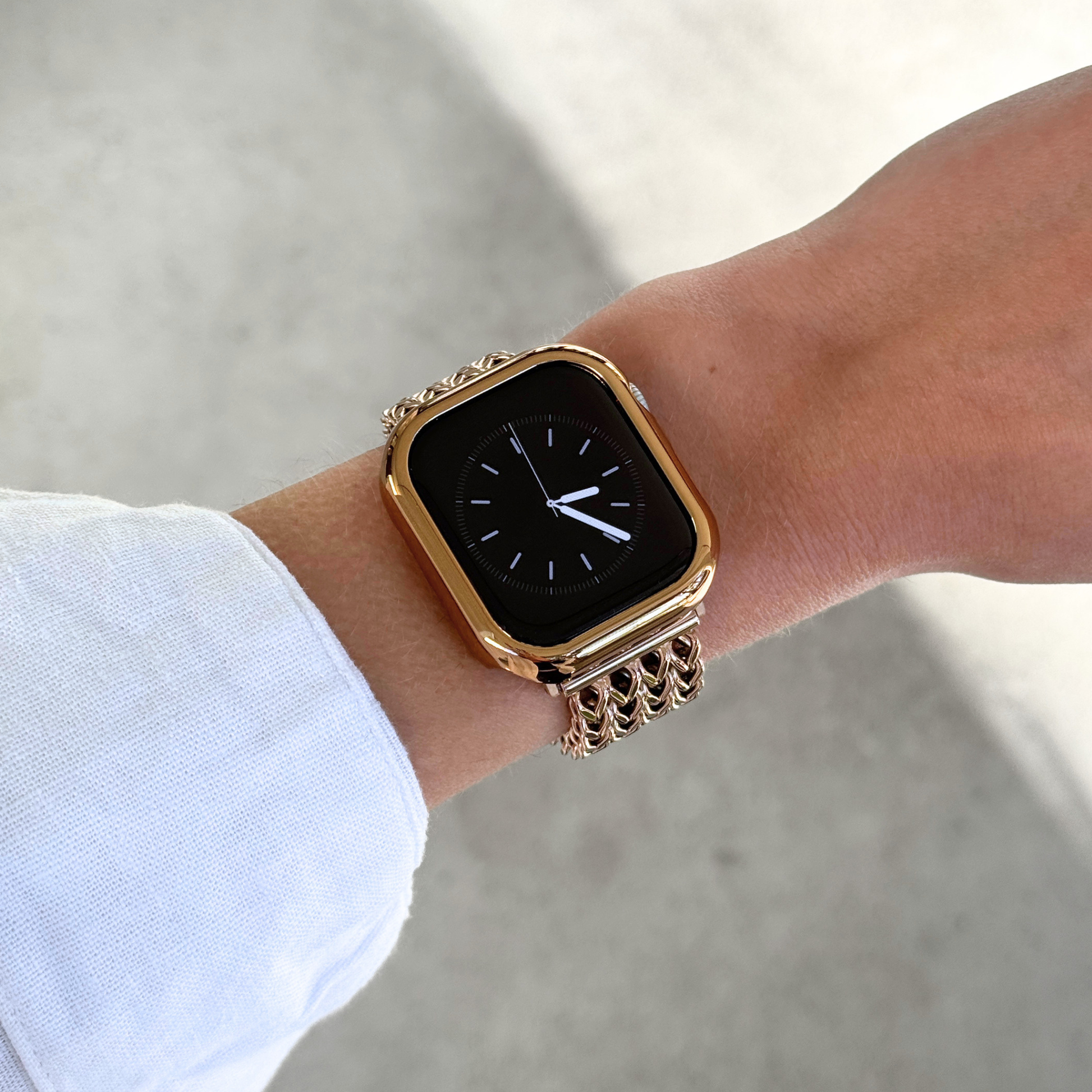 A close-up of an Apple Watch with a rose gold-toned bezel, bumper case cover, with a matching chain-link band worn on a person's wrist, set against a soft gray concrete background.