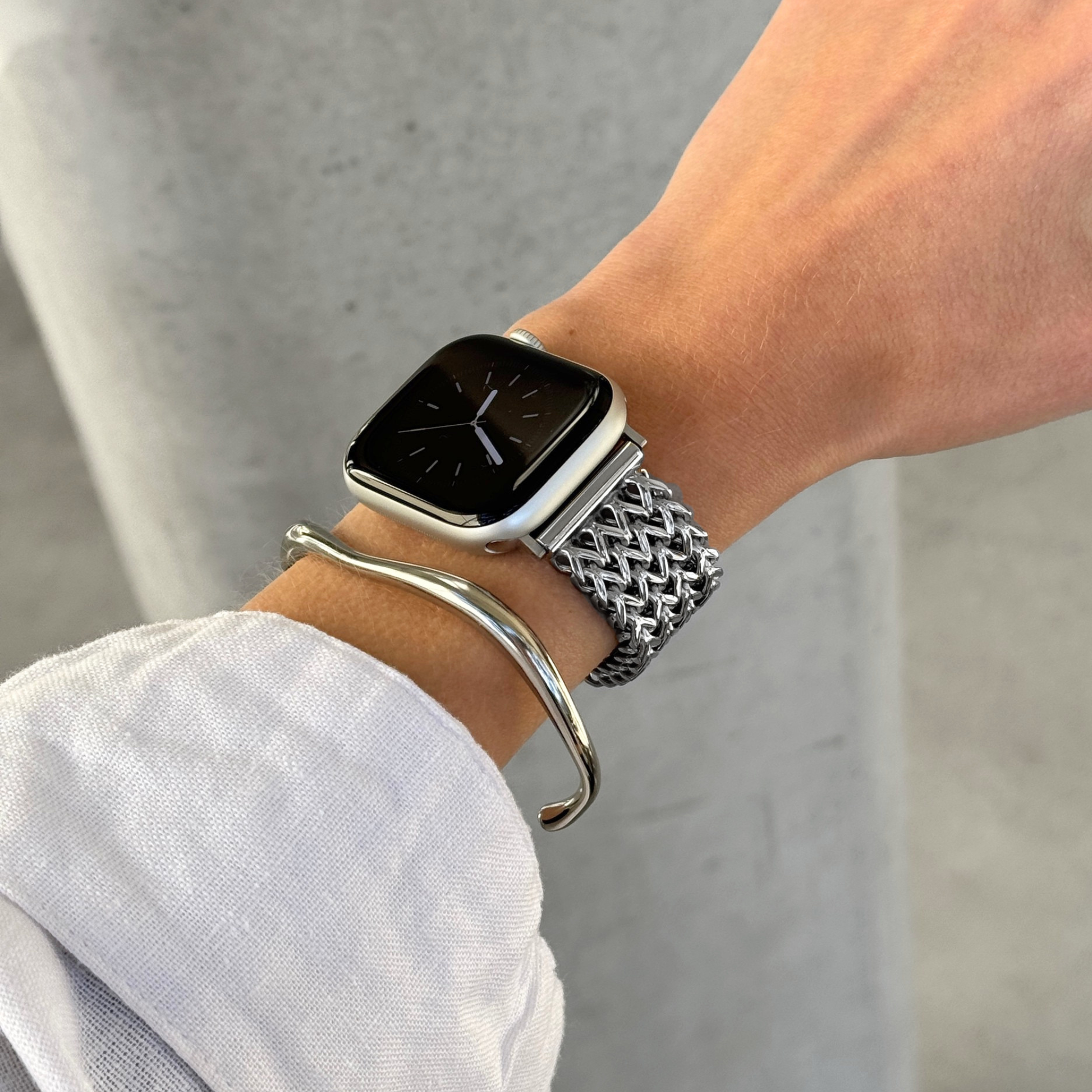 A close-up of a person's wrist wearing an Apple smartwatch with a silver mesh band and a silver curved bangle bracelet, against a light gray background.