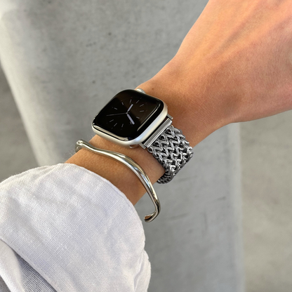 A close-up of a person's wrist wearing an Apple smartwatch with a silver mesh band and a silver curved bangle bracelet, against a light gray background.