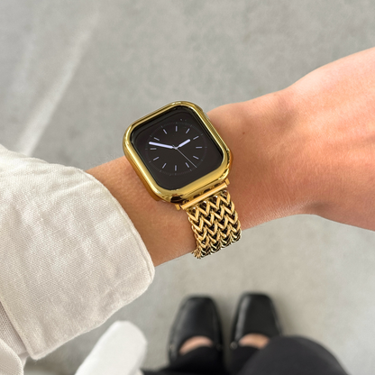 Apple watch with a gold bumper case and a woven metallic gold band, shown worn on a person's wrist against a light gray floor background.