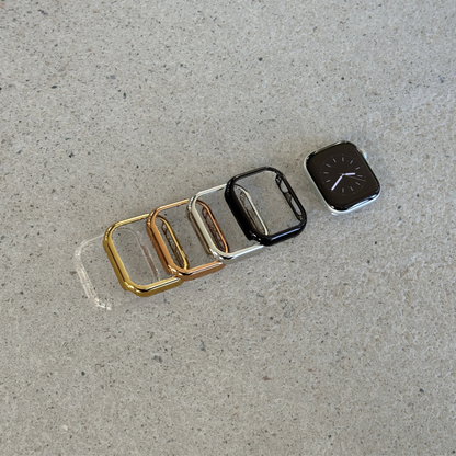 Five smartwatch bezel cases in gold, rose gold, silver, and black are arranged in a row on a textured gray concrete surface, with one Apple watch shown next to the black case.