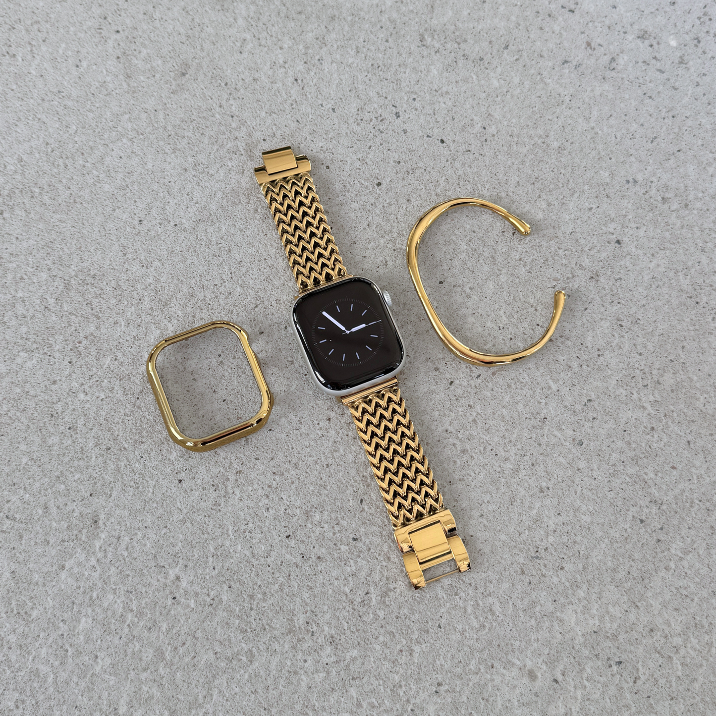 Gold-tone metal Apple watch band, matching watch case and cuff bracelet. Components arranged on a light gray textured surface. The band has a woven zigzag pattern and includes a watch face with a dark display.