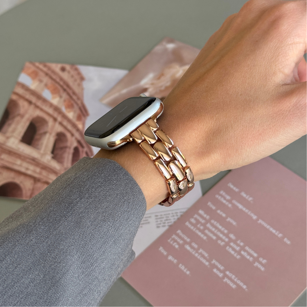 Rose gold slim link Apple Watch band worn with a grey sleeve over a travel magazine, metal strap for Apple Watch 38mm to 49mm
