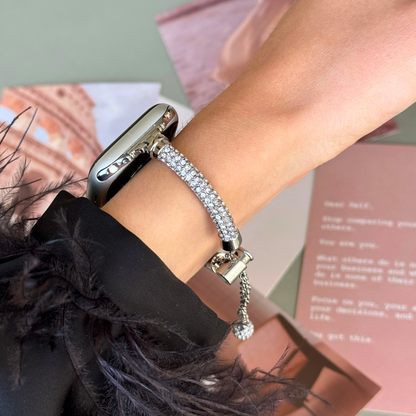 Silver rhinestone Apple Watch band worn on a wrist above pink and green papers, metal strap for Apple Watch 38mm to 49mm