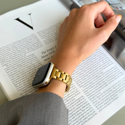 Gold slim link Apple Watch band worn on a raised wrist above an open book, stainless steel strap for Apple Watch 38mm to 49mm