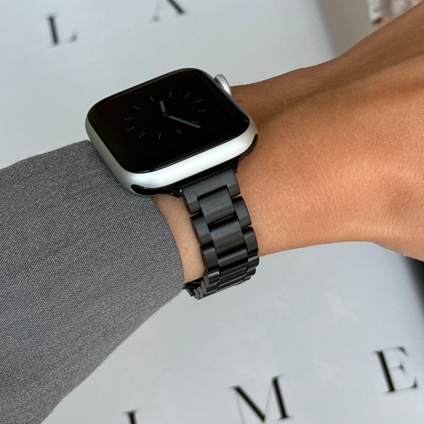 Black slim link Apple Watch band worn with a grey sleeve beside a magazine page, stainless steel bracelet for Apple Watch 38mm to 49mm