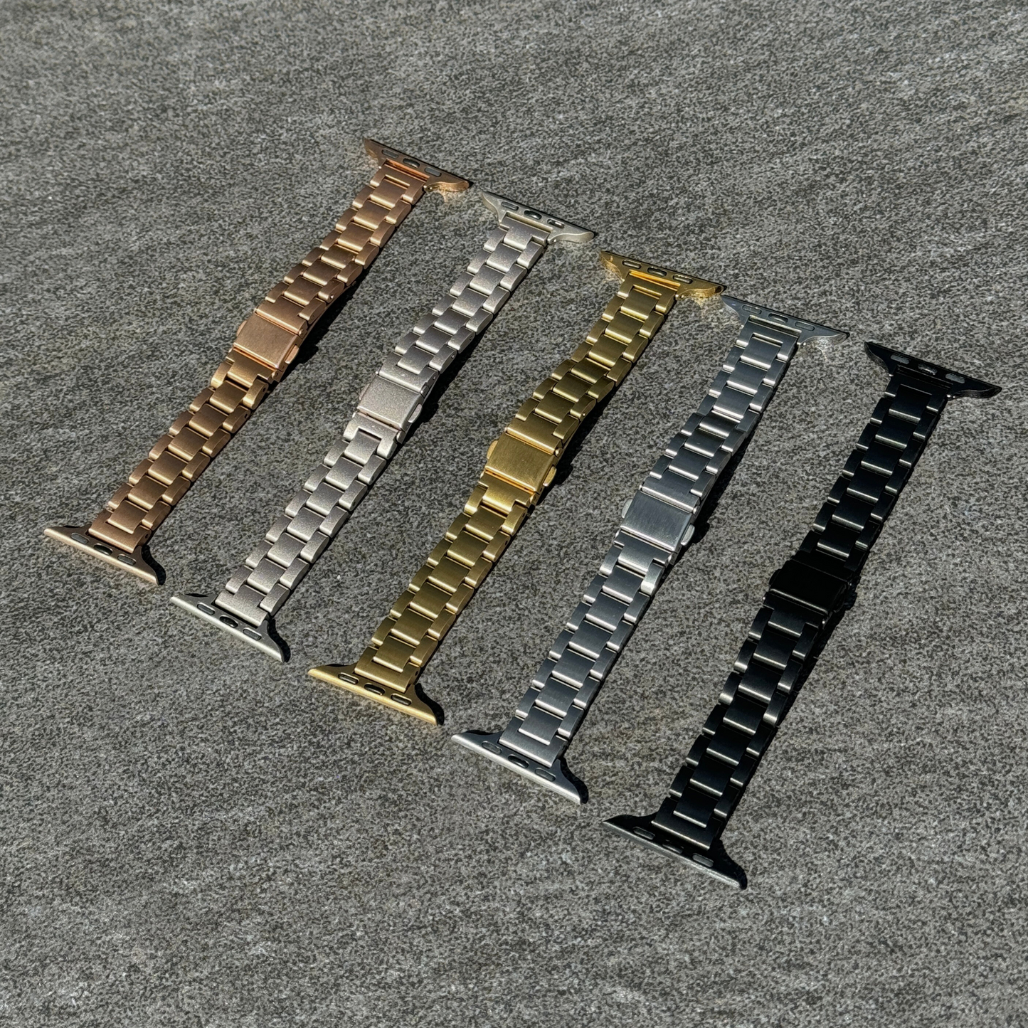 Rose gold, starlight, gold, silver and black slim link Apple Watch bands laid out on grey stone, stainless steel straps for Apple Watch 38mm to 49mm