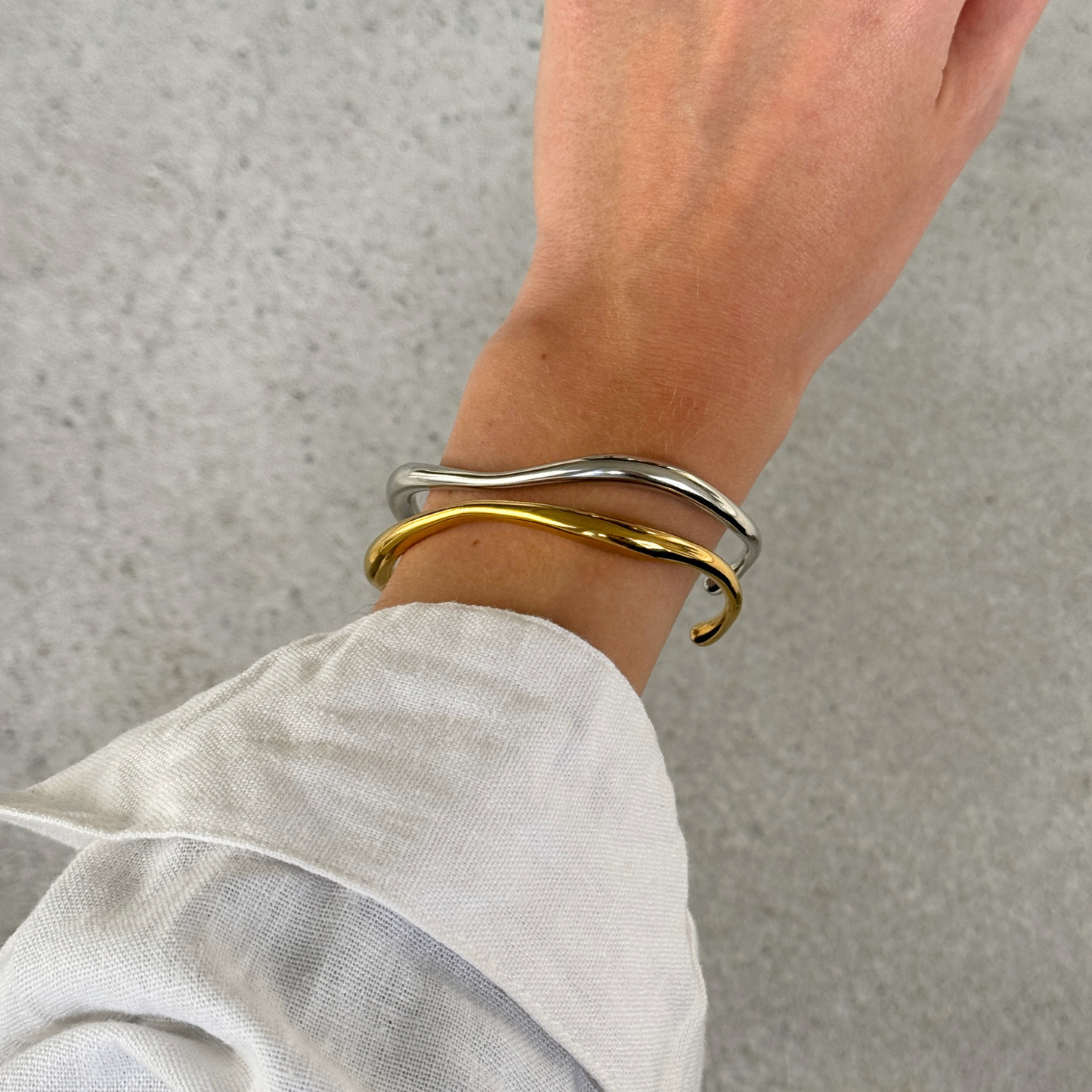 Two thin wavy cuff bracelets, one silver and one gold, worn on a person's wrist with a white linen sleeve visible.