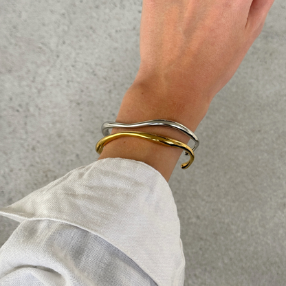 Two thin wavy cuff bracelets, one silver and one gold, worn on a person's wrist with a white linen sleeve visible.
