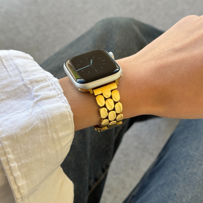 Smartwatch with a gold band on a wrist, wearing a white shirt and gray pants.