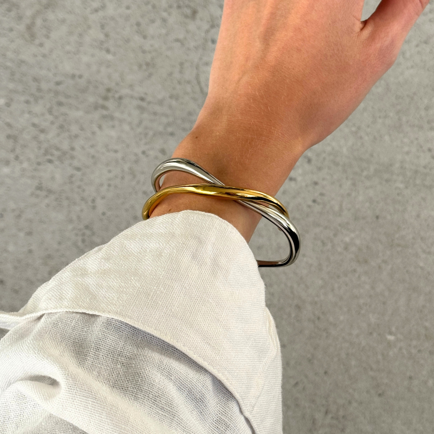Two twisted metal cuff bracelets bangles, one silver and one gold, worn on a person's wrist with a white linen shirt cuff visible, set against a gray concrete background.