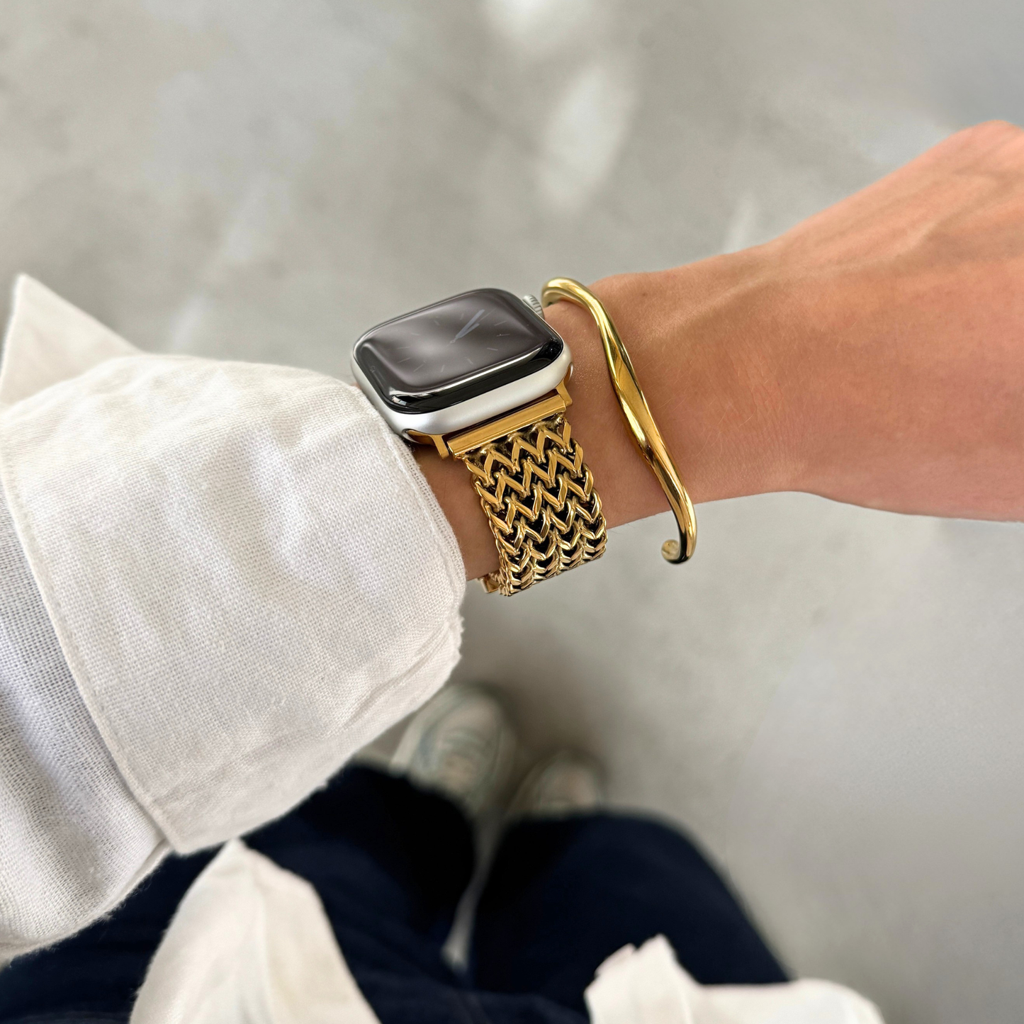 A close-up of a person's wrist wearing a gold-toned mesh Apple watch band, along with a gold bangle bracelet and a white shirt.