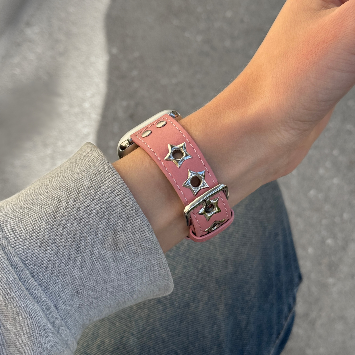 A close-up of a pink leather Apple watch band with silver-tone metal studs and star-shaped hardware, worn on a person's wrist.