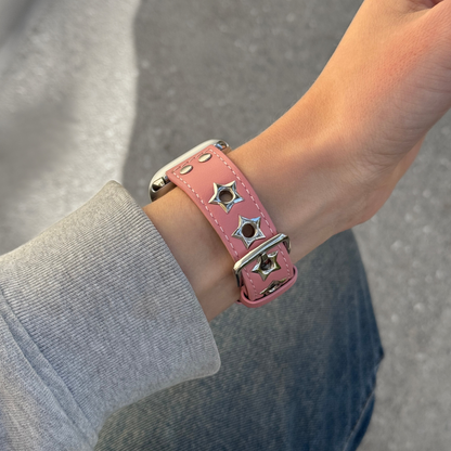 A close-up of a pink leather Apple watch band with silver-tone metal studs and star-shaped hardware, worn on a person's wrist.