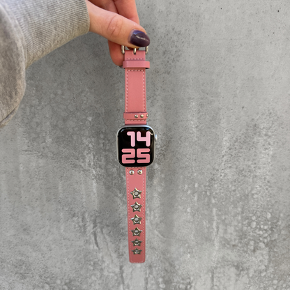 A pink leather Apple watch band with silver-tone star studs, held by a hand against a textured gray concrete wall.