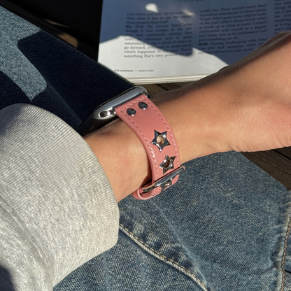 A close-up lifestyle shot of a person wearing a pink leather watch strap with star-shaped metal studs on an Apple Watch. The person is wearing a gray long-sleeve shirt and blue jeans, with a book in the background.