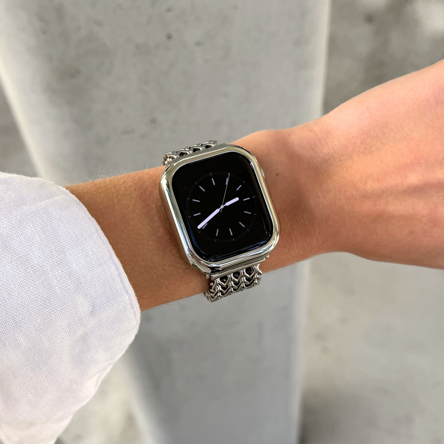 A close-up of a person's wrist wearing a silver-toned metal link Apple watch with a rectangular black screen and a polished silver bezel, set against a blurred gray concrete background.