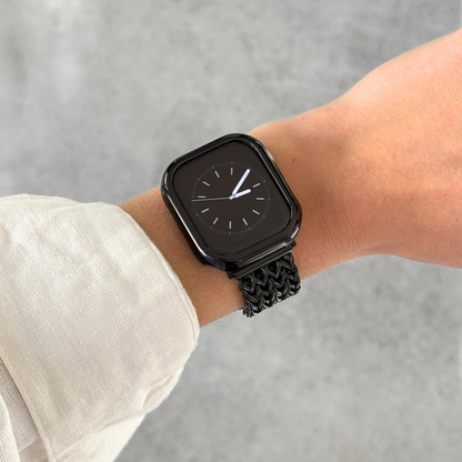 A close-up of a person's wrist wearing a black Apple smartwatch with a black mesh band and a black metallic bumper case cover, against a light gray textured background.